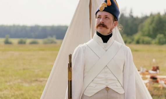 A historical reenactor in Napoleonic military uniform standing by a tent in open countryside.