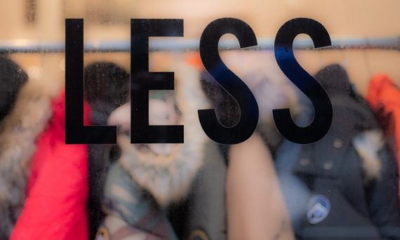The word “less” on a window, in focus with clothes behind it. Commentary on consumerism and waste in the fashion industry.
