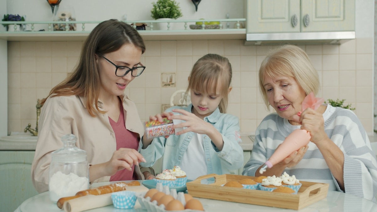 Happy family kid, young mom and senior granny are decorating cupcakes busy with culinary activity in kitchen. Apartment, people and cookery concept.